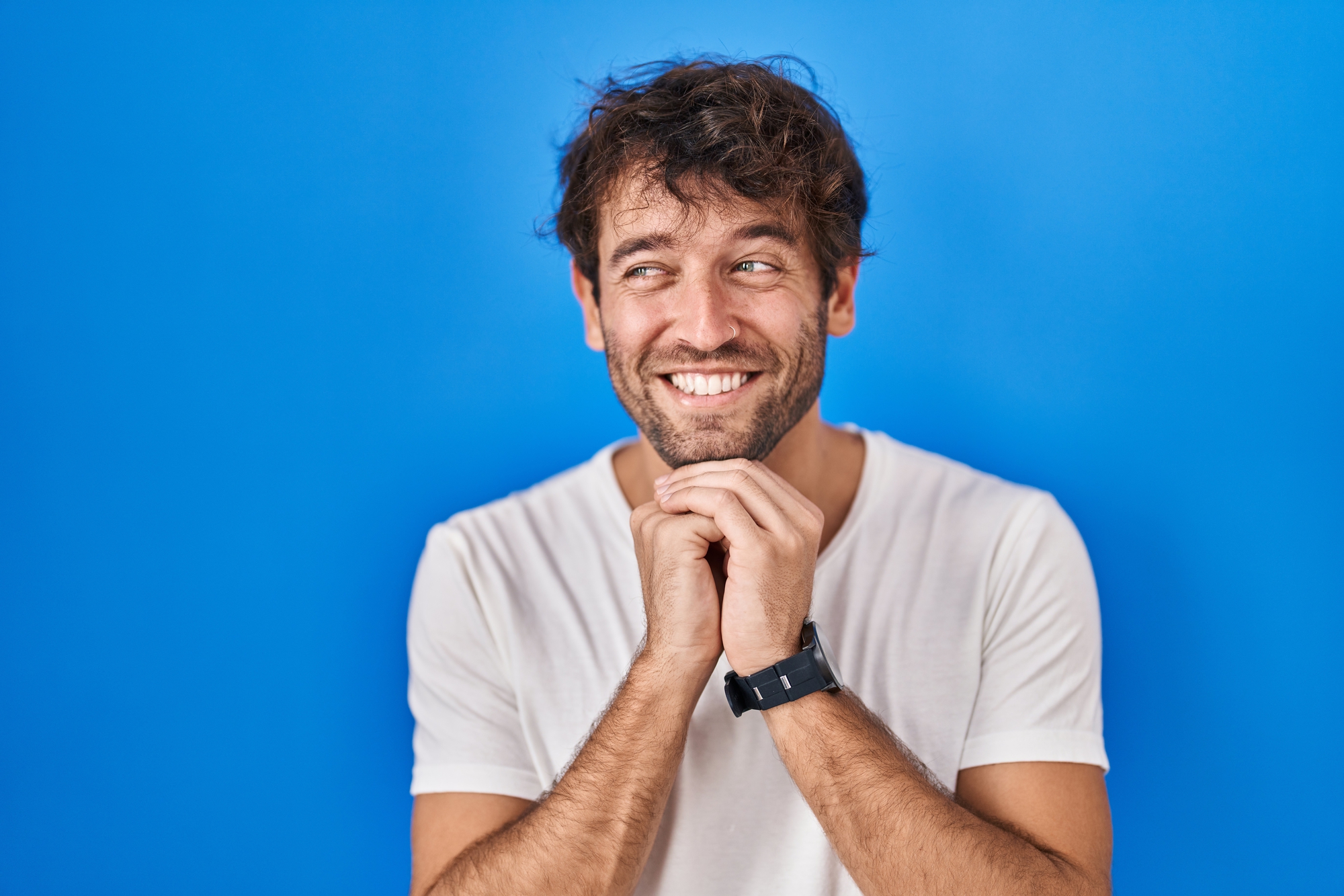 Hispanic young man standing over blue background laughing nervous and excited with hands on chin looking to the side