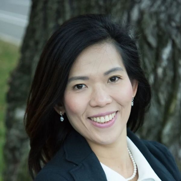 Head-and-shoulders portrait of a smiling Asian woman in a blazer, outdoors with trees in the background (professional headshot).